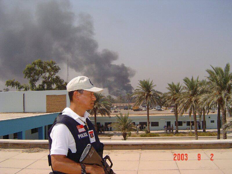 A man wearing a white cap and navy blue vest that says POLICE on it, with a Singapore flag. He is holding a rifle. In the background, there is a row of palm trees and smoke billowing into the sky in the distance.