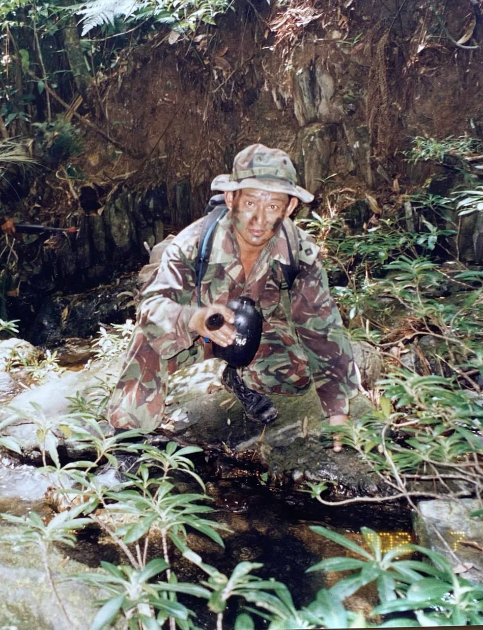 A soldier dressed in camouflage, holding a water canteen, in the middle of the jungle.