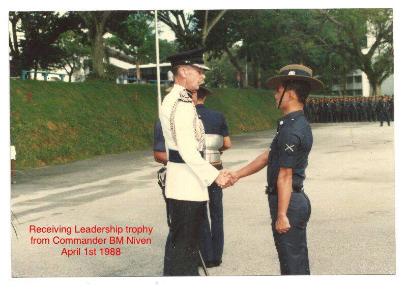 An officer dressed in a white coat shakes the hand of a younger man, dressed in a navy blue police uniform. There is text on the photo that reads, "Receiving leadership trophy from Commander BM Niven, April 1st 1988."