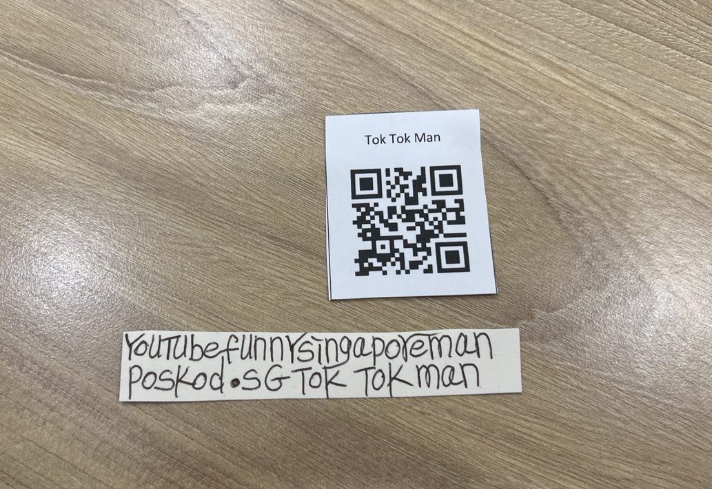 Two white slips of paper on a table. One has a QR code labelled "Tok Tok Man". The other has "Youtube Funny Singapore Man / poskod [sic] / SG Tok Tok Man" written on it.