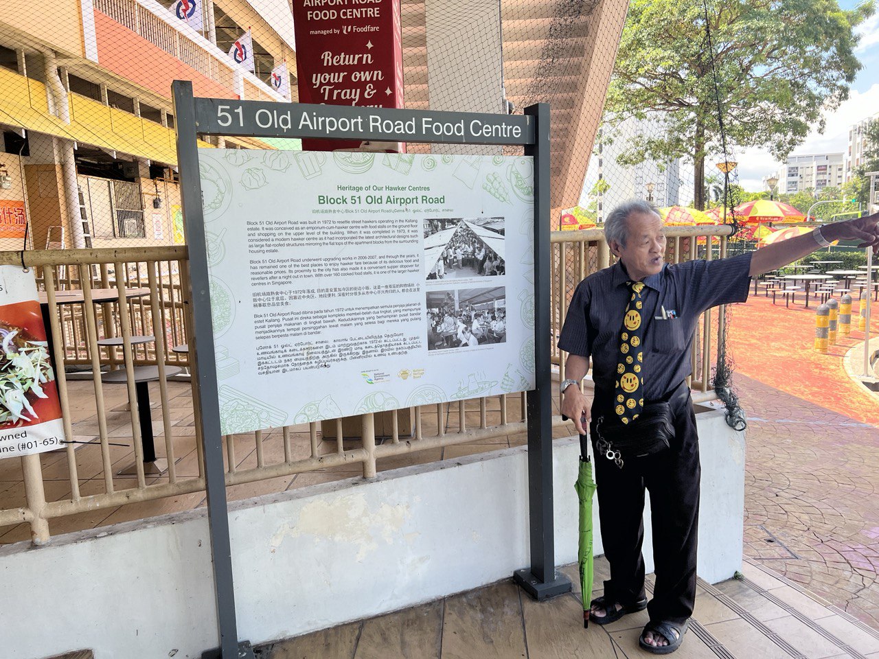 Lai standing next to a sign at Old Airport Road Food Centre, pointing towards the right side of the image.