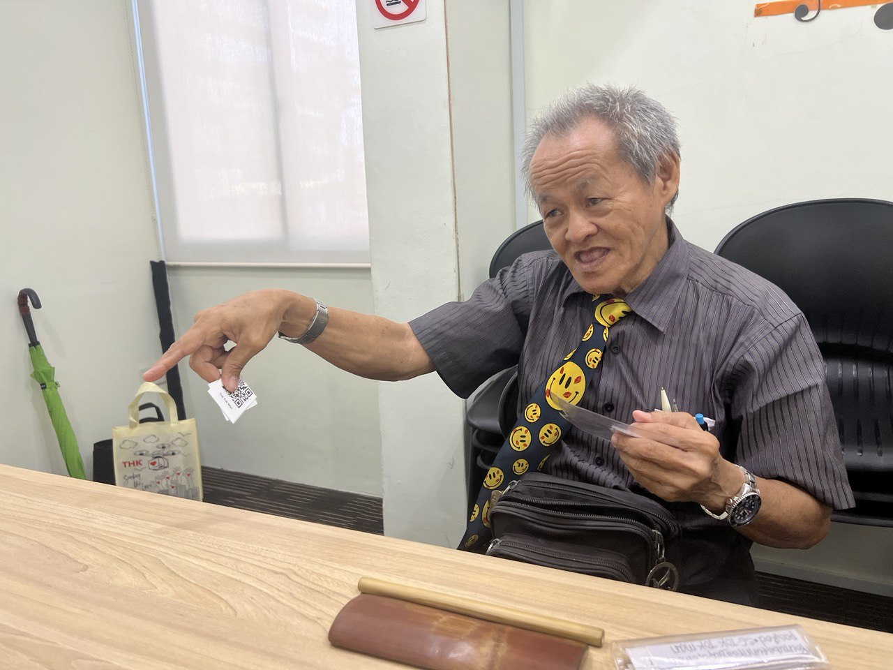 Lai sits on a chair in front of a desk, holding some slips of paper in his hand. He is dressed in a grey collared shirt, along with a dark blue tie that has yellow smiley faces and red cartoon lipstick stains over it. He also has one wristwatch on each hand.
