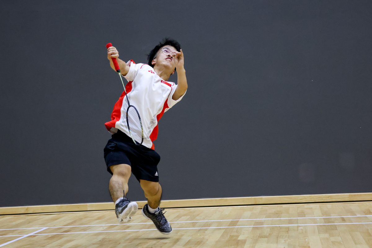 Lim is dressed in a white t-shirt and black shorts. He jumps upwards and prepares to hit the shuttle with his racket.