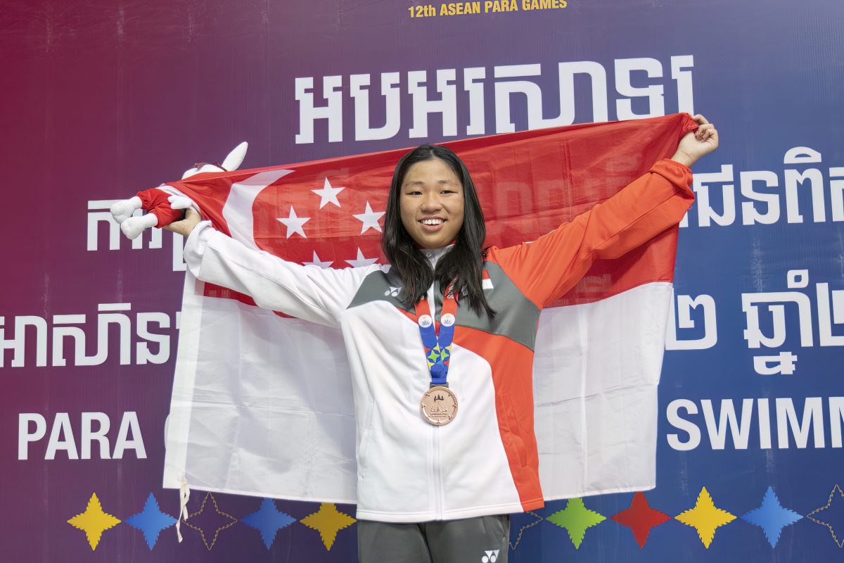 Tong holds a Singapore flag up behind her back, donning a bronze medal and smiling at the camera. She is also wearing a white Team Singapore outfit.