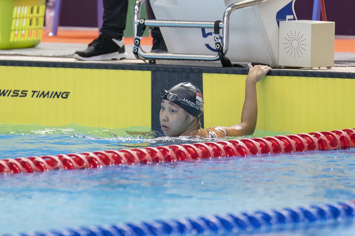 Tong, donning a black swimming cap, holds on to the edge of the pool and checks the scoreboard (offscreen).