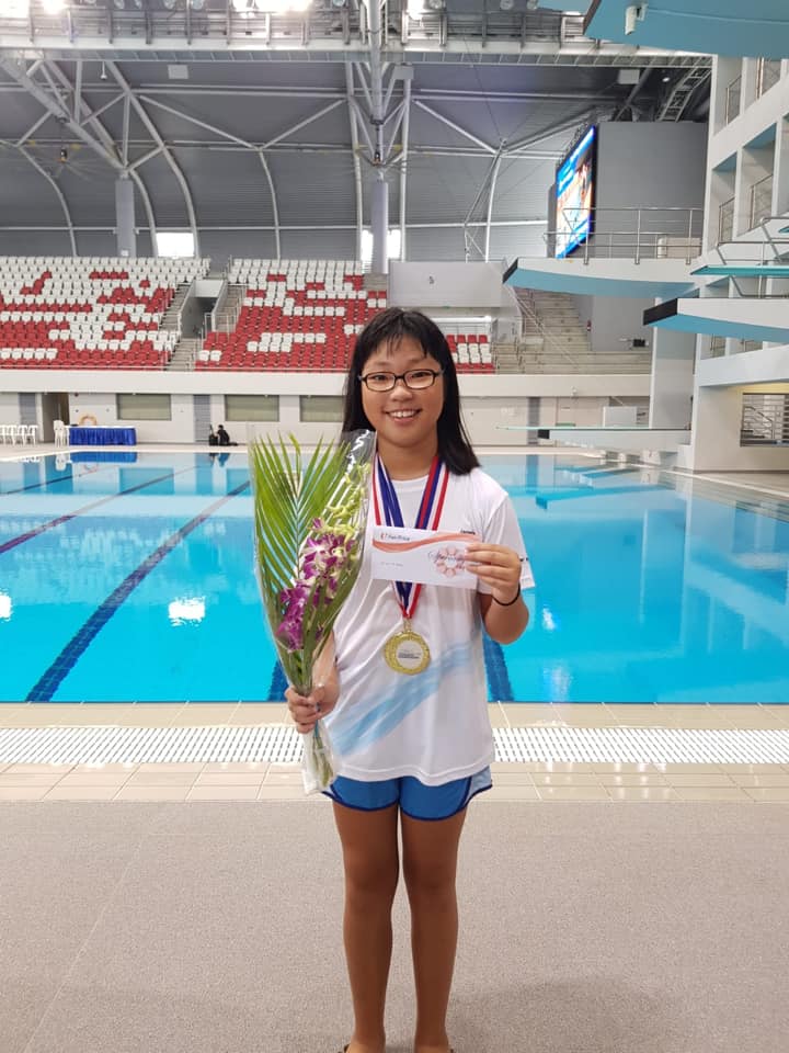 Tong dresses in a white t-shirt, wearing a gold medal and holding a bouquet. She stands in front of the swimming pool and poses for the camera.