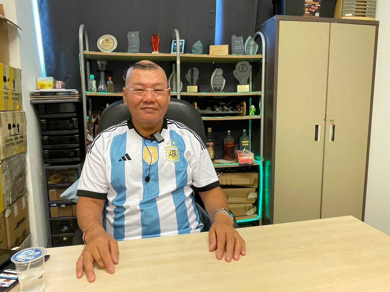 Yu is dressed in a blue-and-white striped shirt. He sits at a table and smiles at the camera. Behind him, there is a shelf and cupboard full of awards, bottles, and other items.