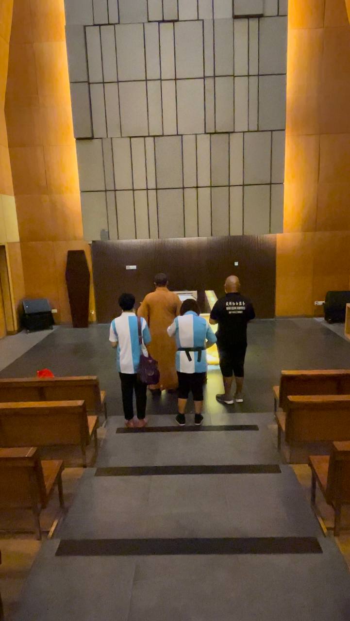 The back view of a monk dressed in brown robes, two people dressed in blue and white shirts, and Yu dressed in a black shirt. They stand at the front of a funeral hall.