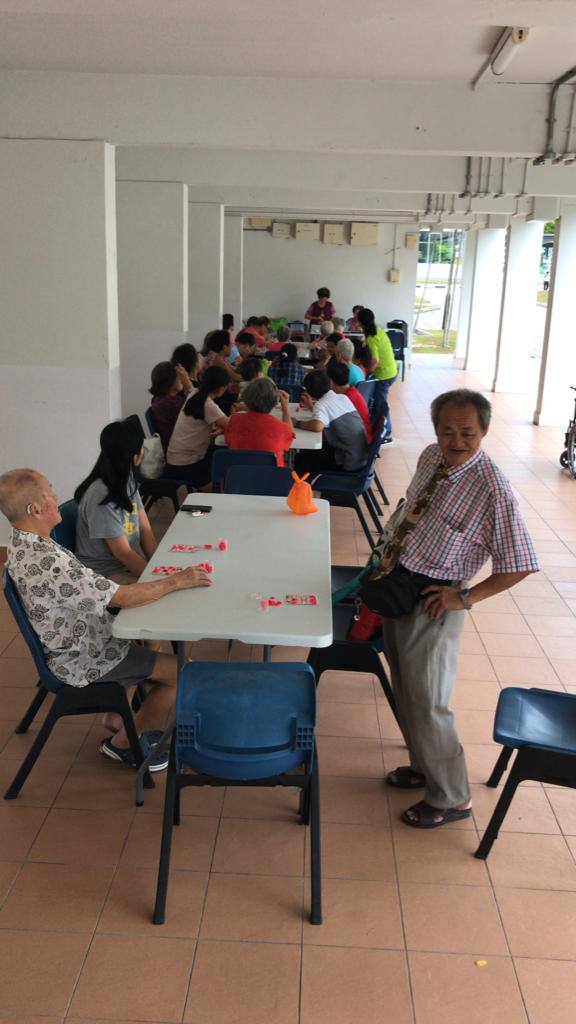 Lai wears a checkered shirt and tie. He is standing at a row of tables in a void deck, together with other elderly folks.