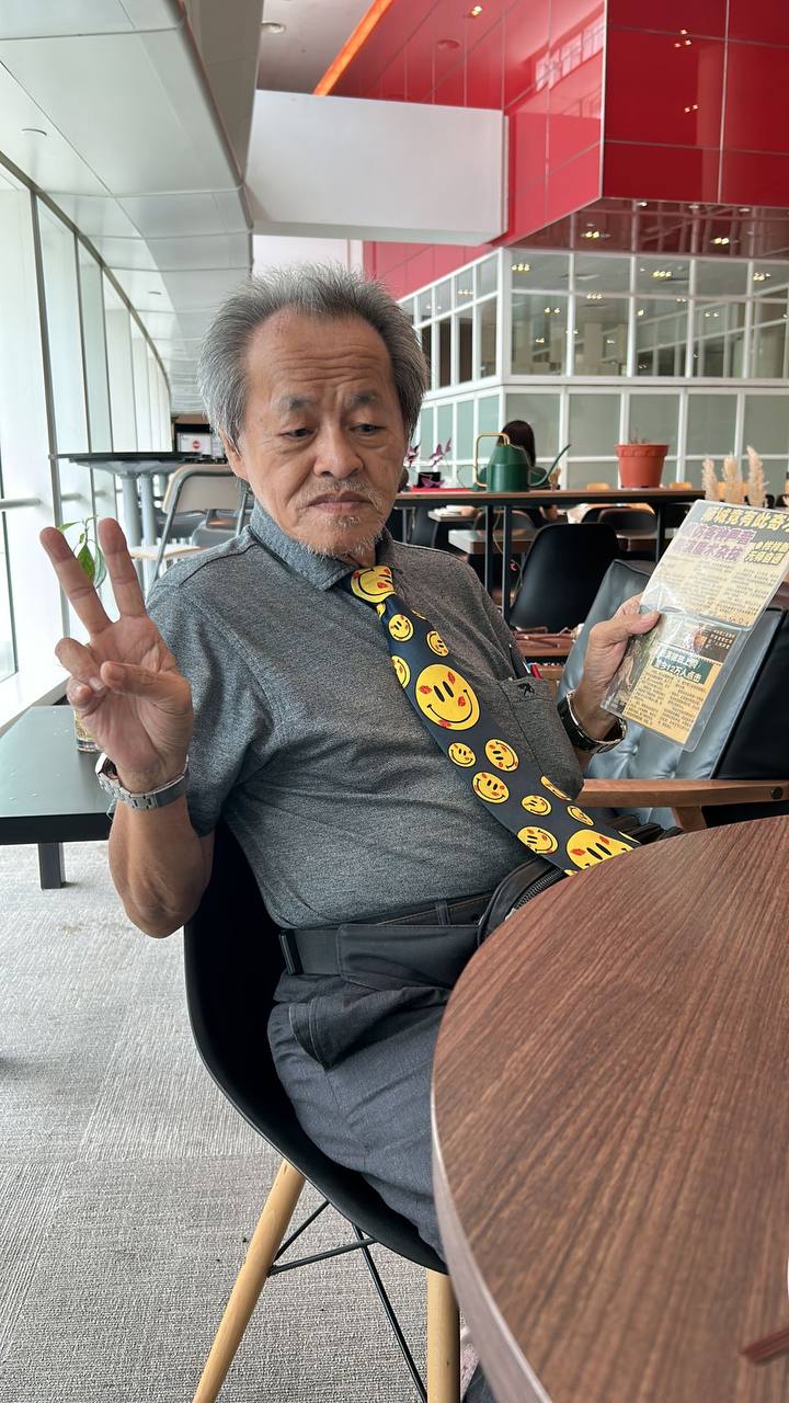 Lai sitting down at a wooden table, holding up a peace sign with one hand, and holding a newspaper clipping in the other hand.