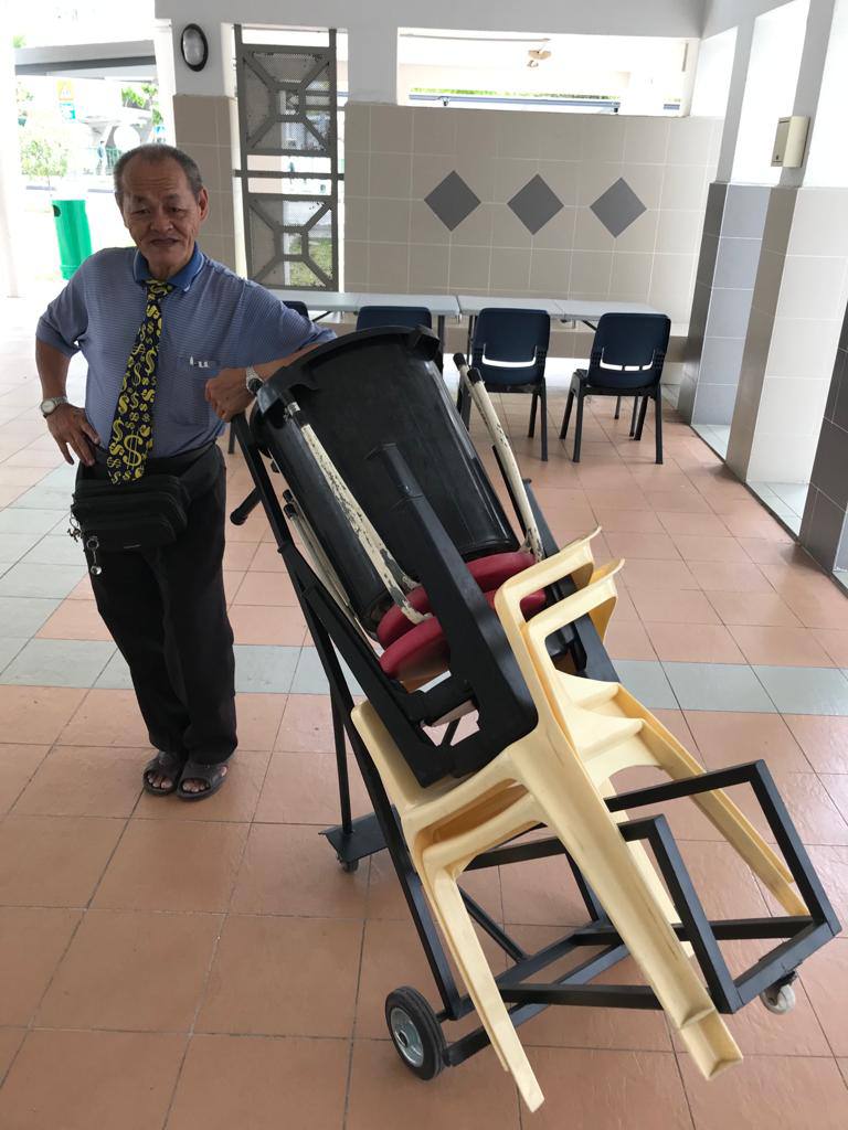 Lai, dressed in a blue shirt and a black tie with fancy yellow symbols, poses beside a stack of chairs that he helped to keep.