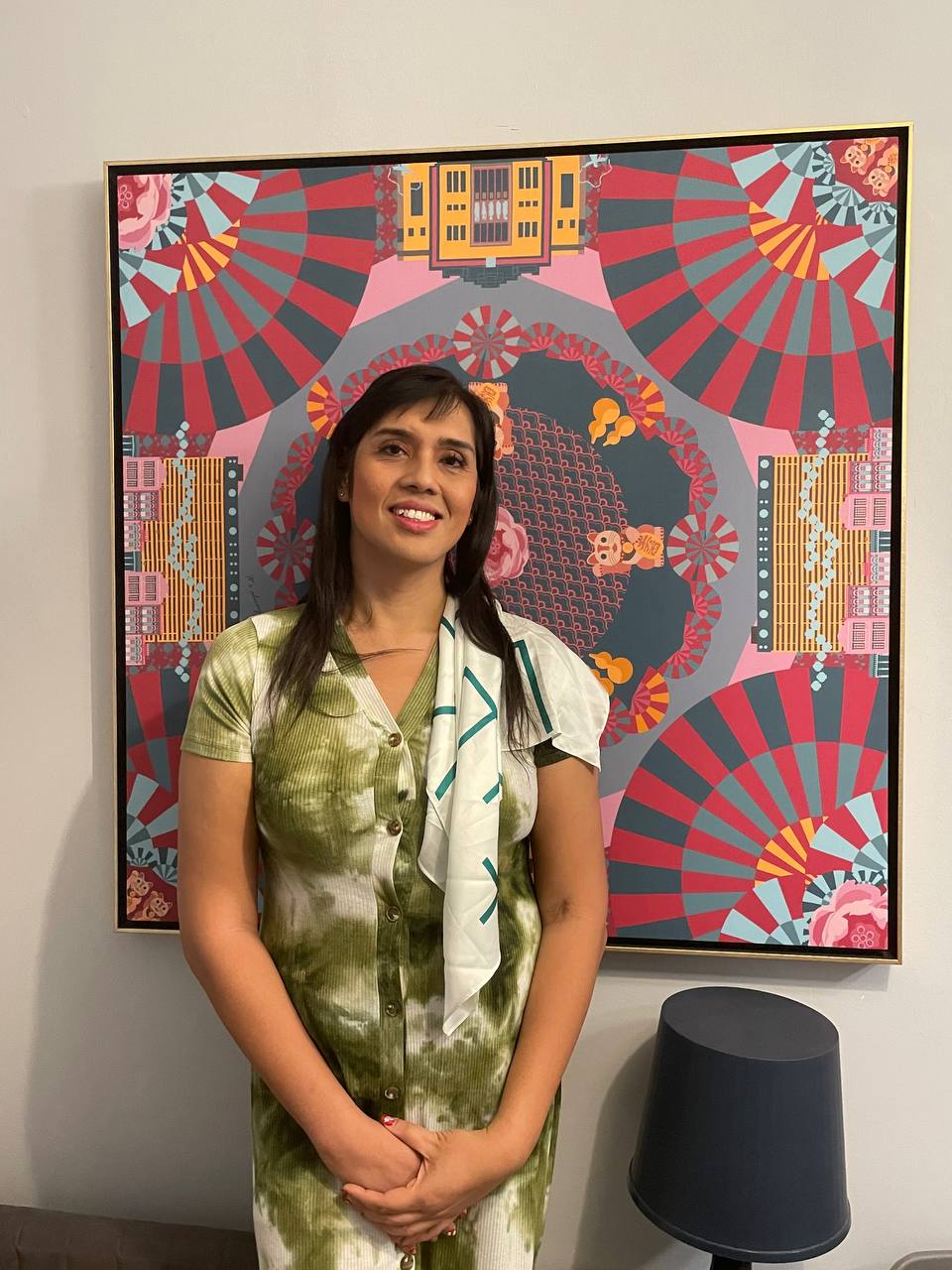Shania dons a white scarf and a green dress. She stands in front of an art piece with pink, blue, and yellow designs on it, and smiles for the camera.