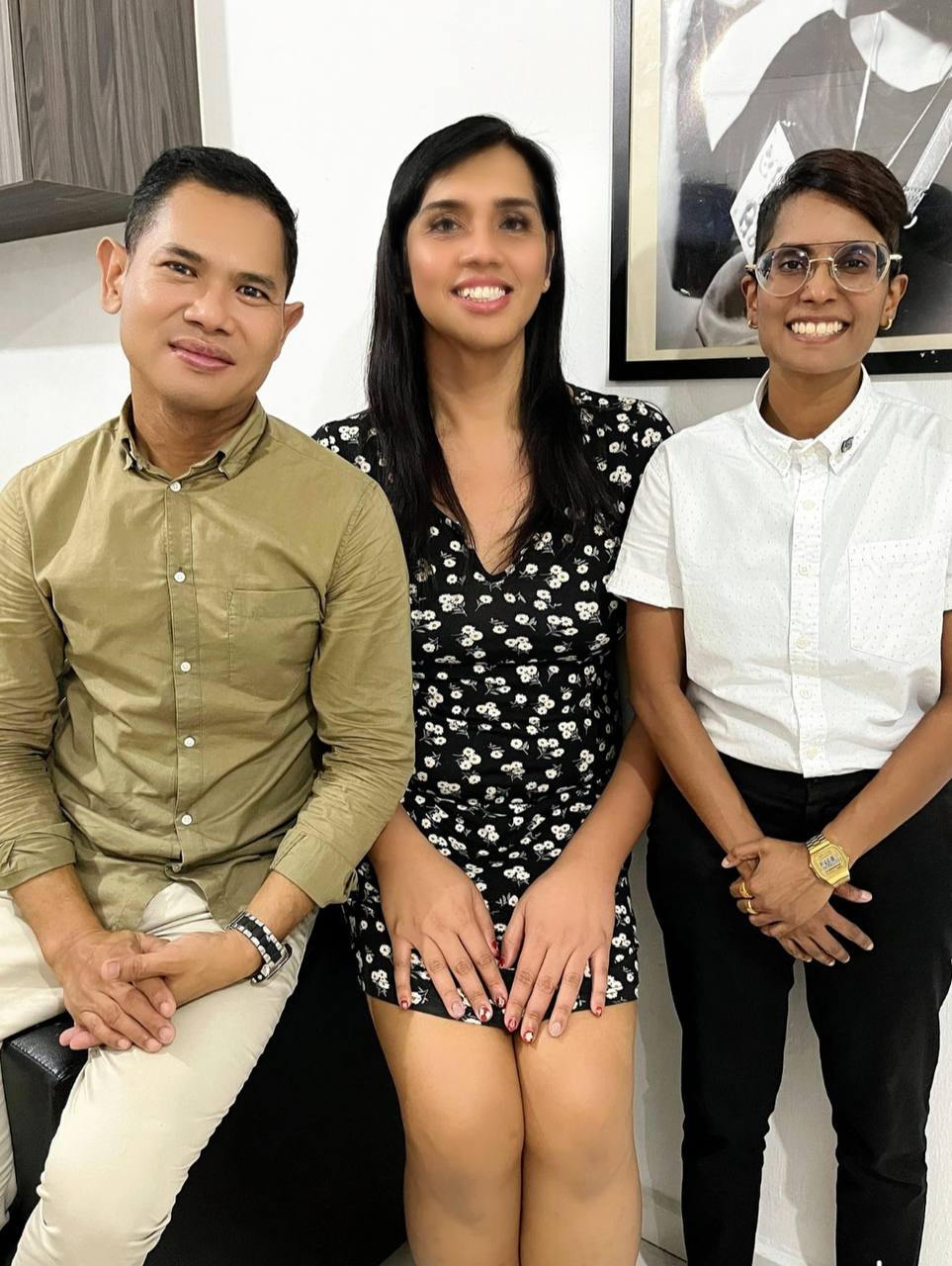 Askre, Shania, and Pereira pose for a photo together. Askre is dressed in a green long-sleeved shirt and khaki pants. Shania dons a black floral dress. Pereira is dressed in a white short-sleeved shirt and black pants.