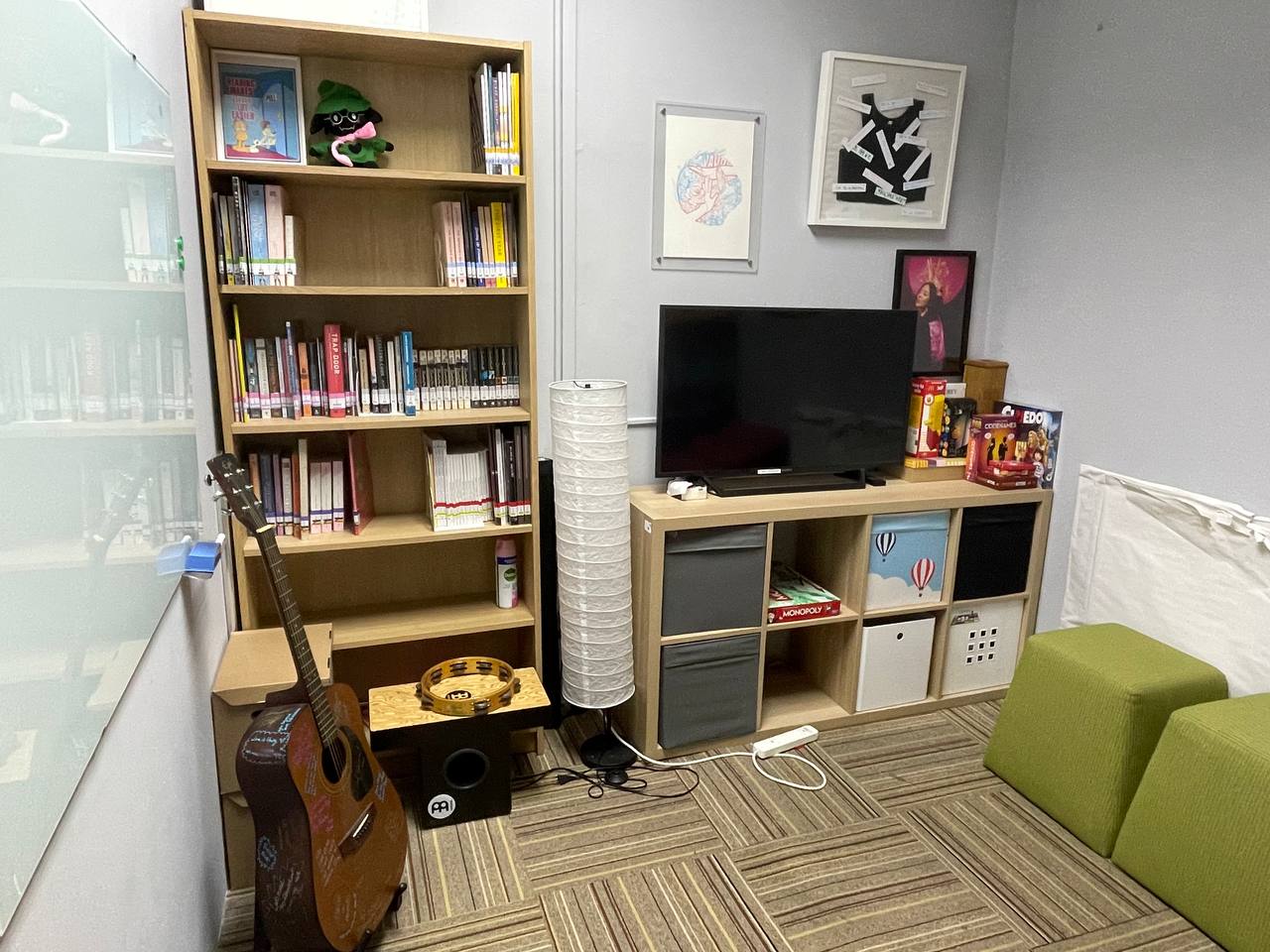A carpeted room with a bookshelf on the left, a guitar sitting on the floor, a television, and some board games.