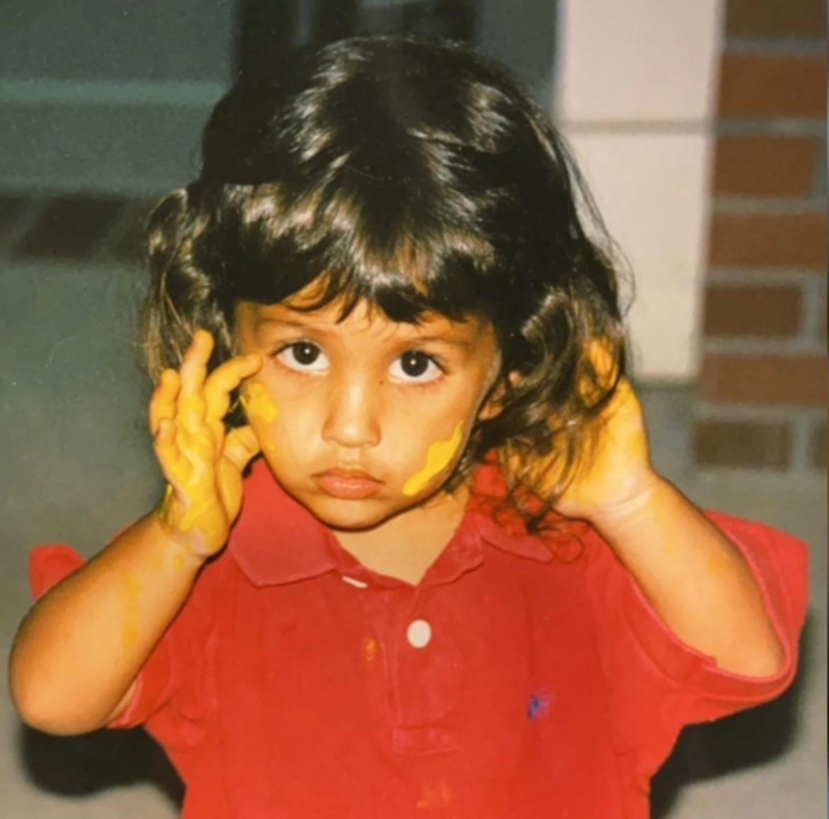 Bailey McNamee as a child wearing a red shirt with yellow paint on her hands/face.